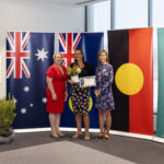 Photo of three women standing and smiling in front of flags. Woman in the middle is holding flowers and a certificate.