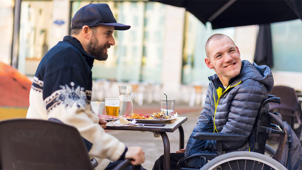 Two men sitting by a coffee table outside. One man is sitting in a wheelchair and looking over his shoulder. He is wearing a blue jacket and smiling. The other man is wearing a blue cap, a white and blue sweater. He is looking at the first man.