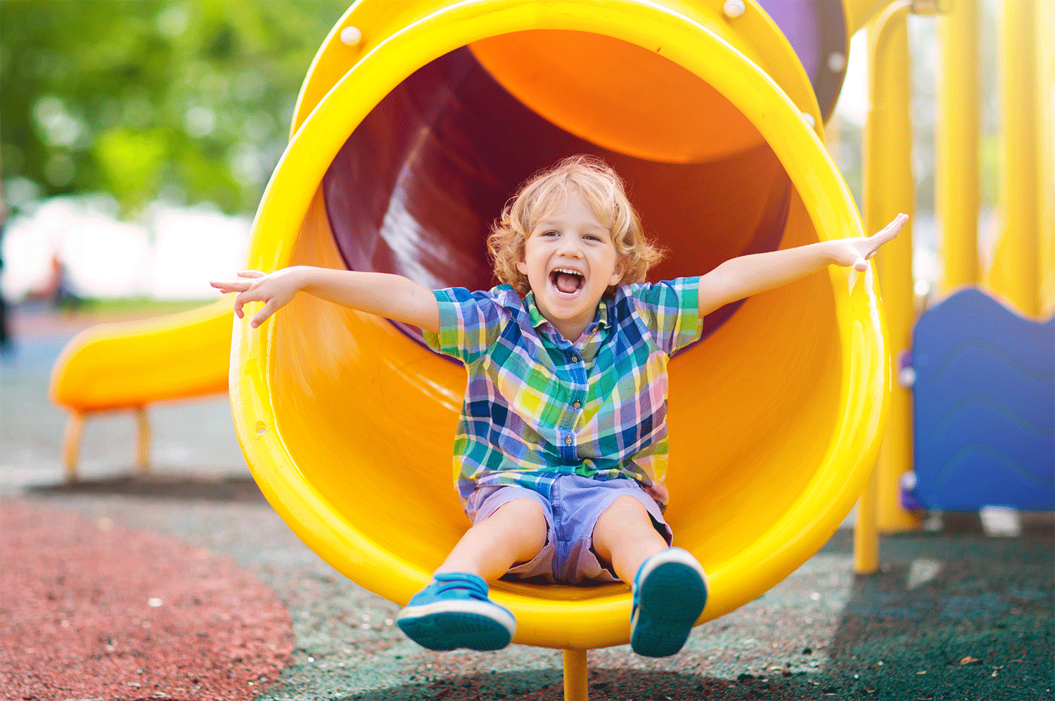 A young, blonde boy is laughing while he is coming down a play ground slide. He has his arms stretched out in joy. he is wearing a colorful checkered shirt and blue shorts.