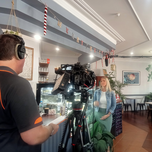 A man with a black shirt and a camera is standing with his back toward the camera. He is filming a blonde woman, wearing a denim jacket and a green skirt. They are in a cafe, and standing by the cafe counter.