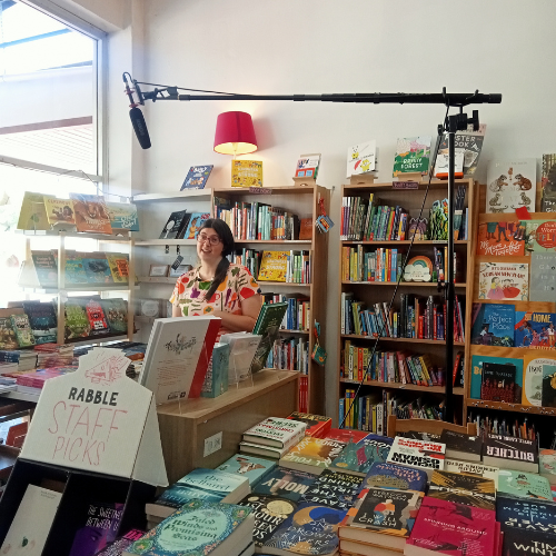A person with dark hair and a colourful dress standing in a book shop. They are surrounded by books. There is a microphone hanging in front of her.