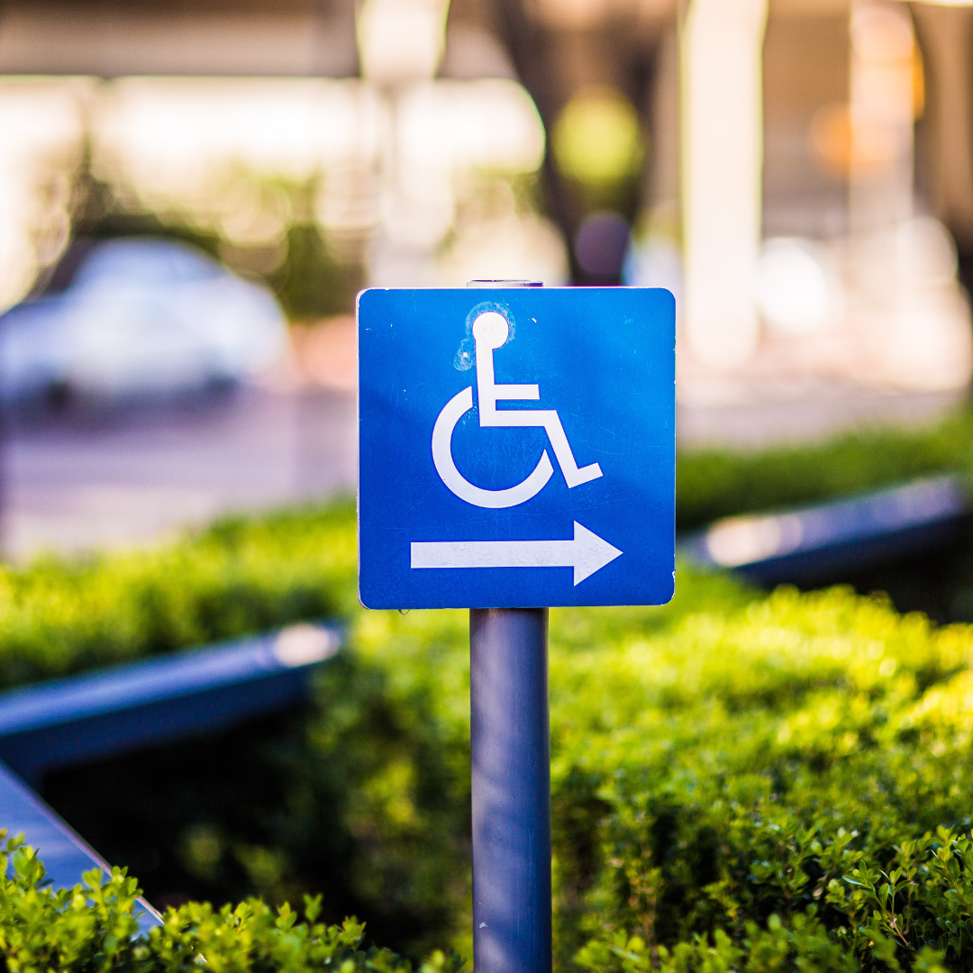A blue sign with a white wheelchair symbol and a right-pointing arrow, indicating accessible access or parking for people with disabilities. The sign is in front of green bushes and a blurred background.