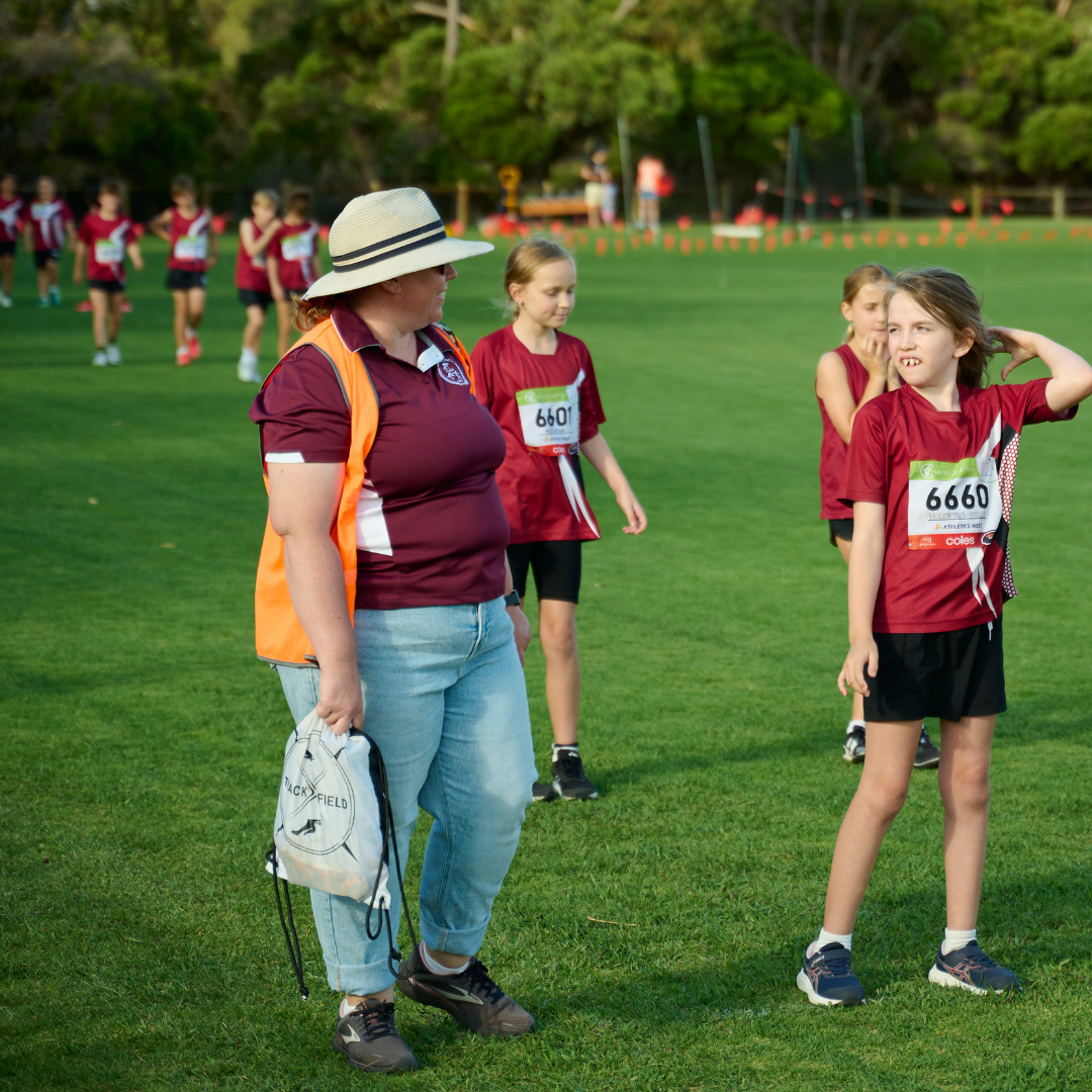 An adult in a hat and orange vest talks to a child wearing a race bib on a grassy field. Other children in matching sportswear are in the background, preparing for a race or event.