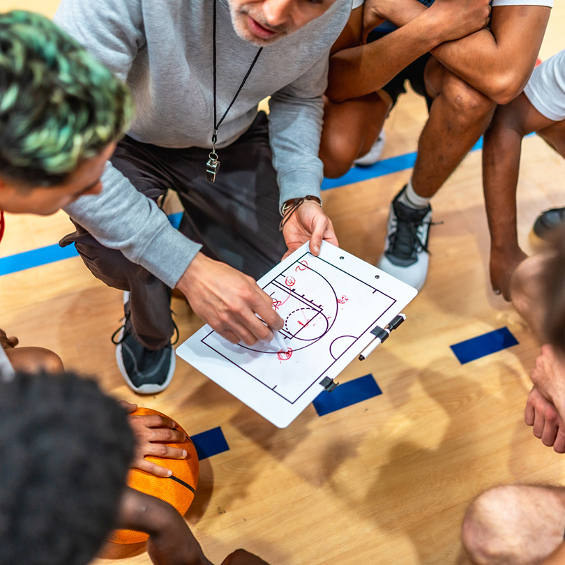 A coach uses a clipboard with a basketball court diagram to discuss strategy with players gathered around on a gym floor.