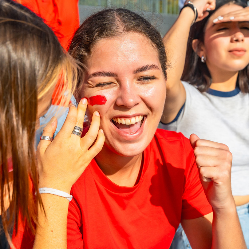 Smiling young woman in a red shirt gets her cheek painted with a red stripe while celebrating outdoors with friends.