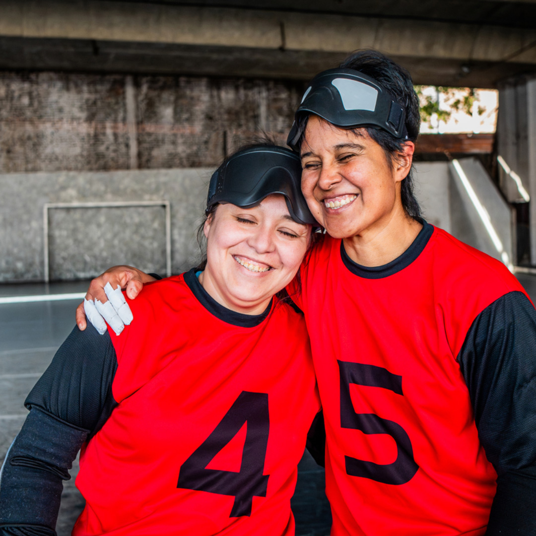 Two smiling women wearing red jerseys with numbers 4 and 5 and black eye masks stand arm in arm, appearing happy and close, in an indoor sports setting.