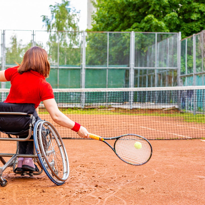 A person in a wheelchair, wearing a red shirt, plays tennis on an outdoor clay court, hitting a yellow tennis ball with a racket. Green trees and a wire fence are in the background.