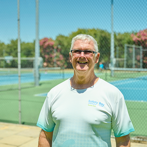 An older man wearing glasses and a Safety Bay Tennis Club shirt smiles while standing in front of outdoor tennis courts on a sunny day.