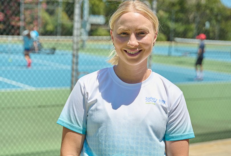 A woman with blonde hair tied back, wearing a white and blue Safety Bay t-shirt and a black skirt, stands smiling in front of a tennis court with players in the background.