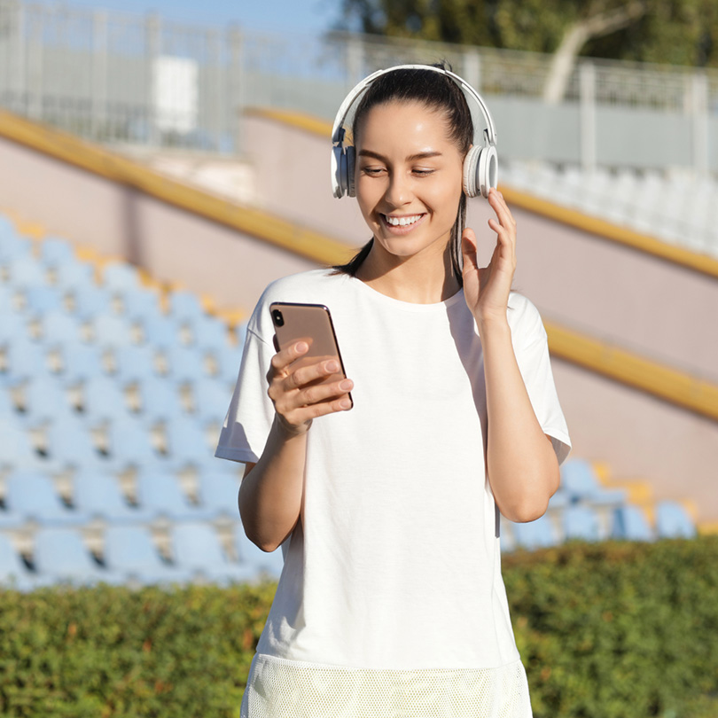 A young woman wearing headphones and a white t-shirt smiles while looking at her smartphone, standing in front of empty stadium seats outdoors.