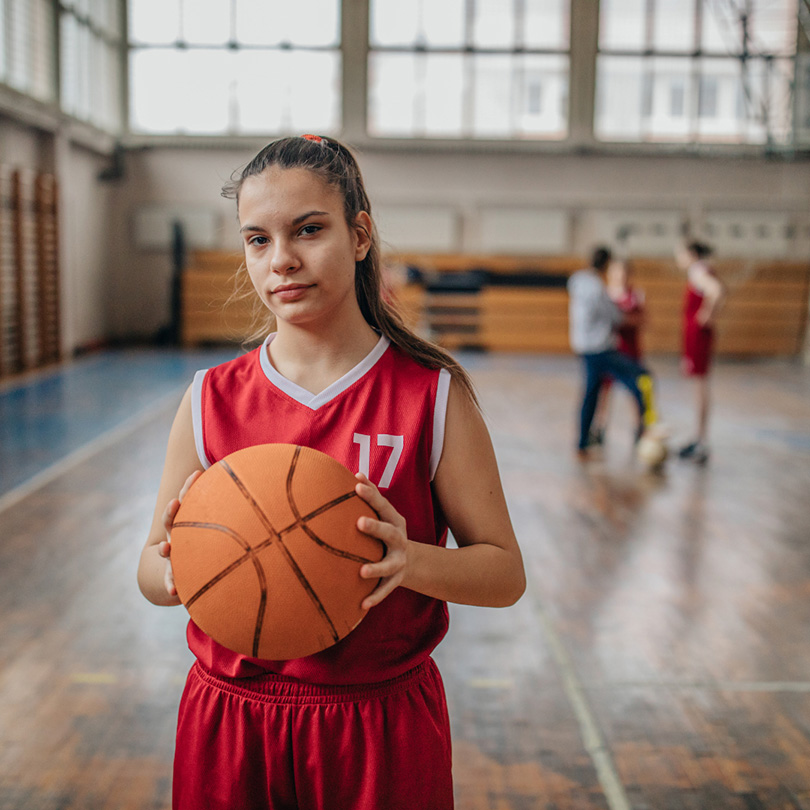 A teenage girl in a red basketball uniform holding a basketball stands in a gymnasium, looking at the camera. Other players in red uniforms are visible in the blurred background.