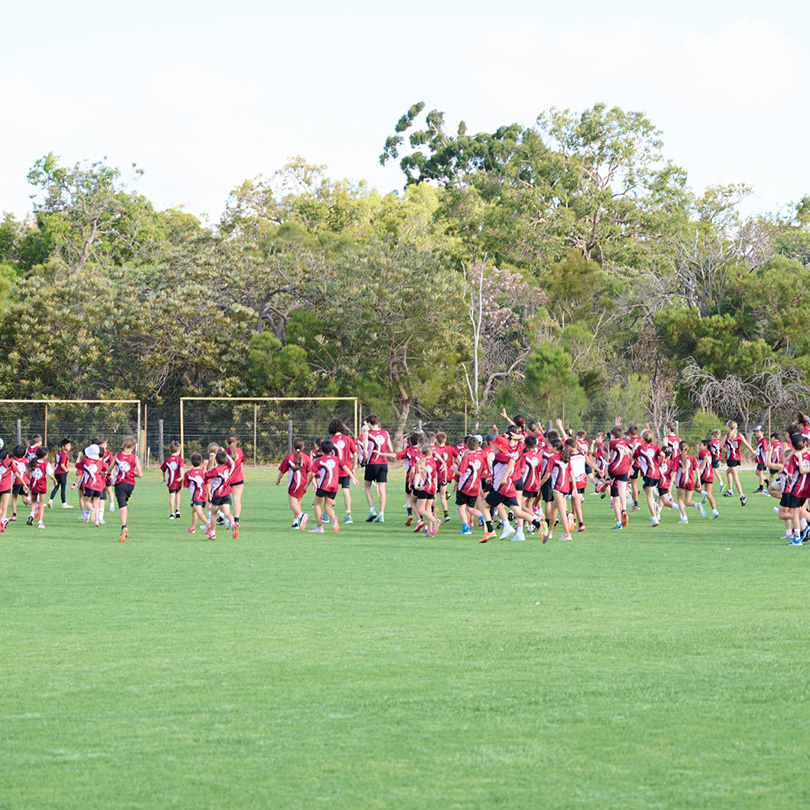 A large group of students in matching red and black uniforms gather and walk across a green sports field, with trees and a blue sky in the background.