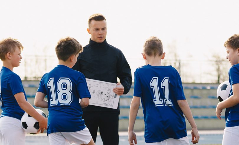 A coach holds a tactic board while giving instructions to four young boys in blue soccer uniforms, each holding a soccer ball, on a field with orange cones.