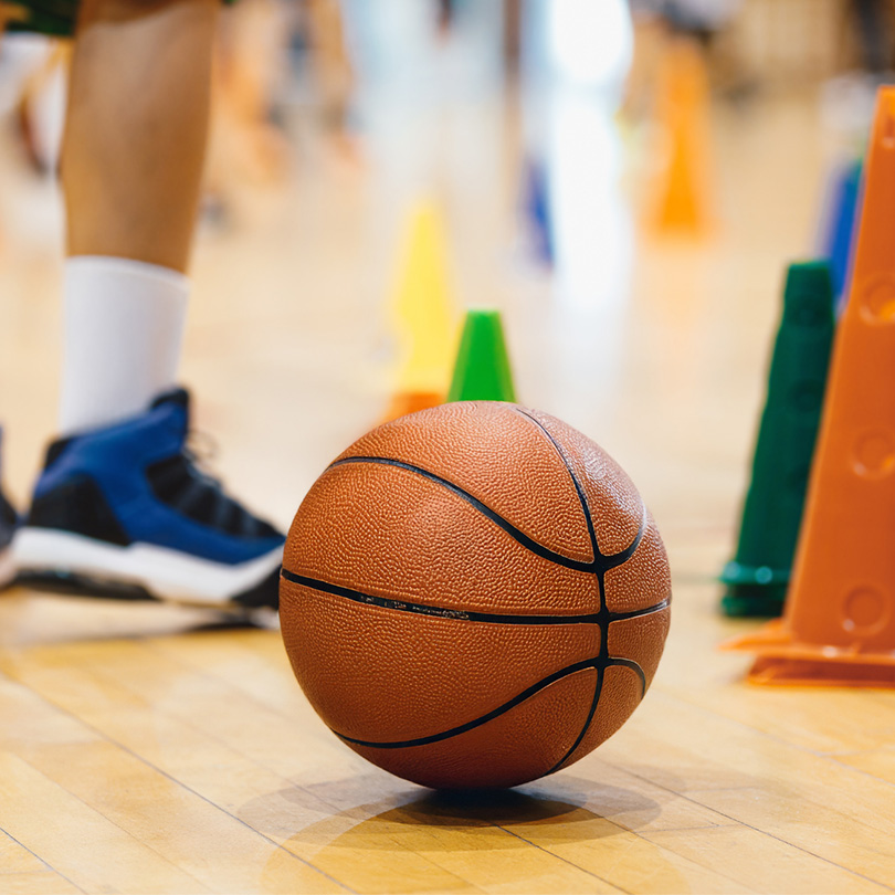 A basketball rests on a polished gym floor near colorful training cones, with a person wearing blue and black athletic shoes standing nearby.