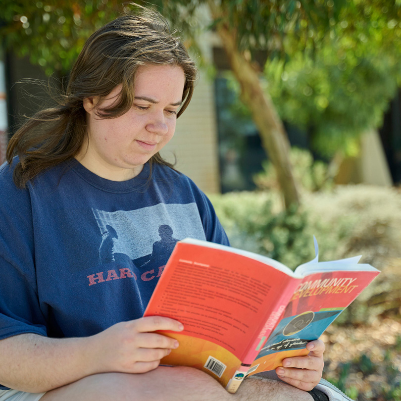 A person with long brown hair sits outdoors reading a book titled Community Development. Trees and a building are visible in the background.