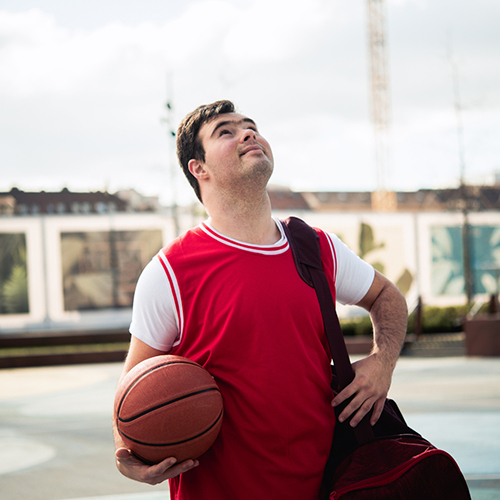 A young man wearing a red basketball jersey and holding a basketball looks up while standing outdoors on a basketball court, with a gym bag slung over his shoulder.