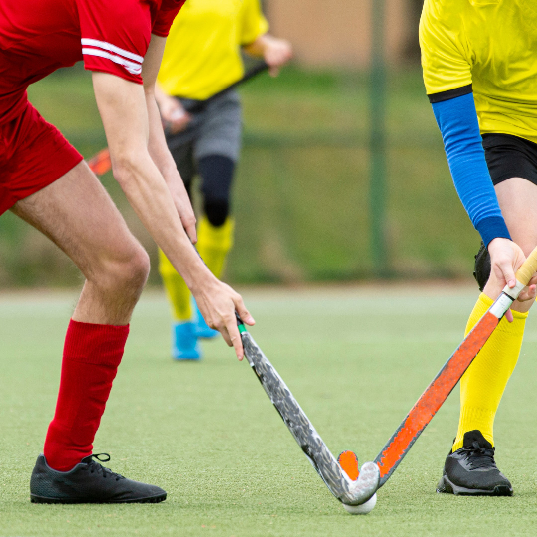 Two field hockey players, one in a red uniform and the other in yellow, compete for the ball on an outdoor field. Only their lower bodies and hockey sticks are visible.