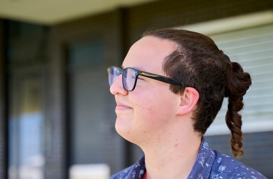 A person with glasses and a ponytail smiles while looking to the side. They wear a red shirt, a patterned blue overshirt, and a button badge, standing outdoors in front of a building with steps and large windows.