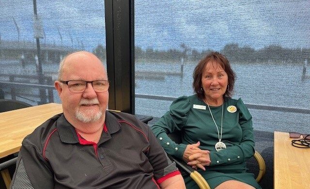 A man and a woman are sitting at a table in a sunlit room. The man wears glasses and a black shirt with red accents. The woman has a green dress, smiling warmly.