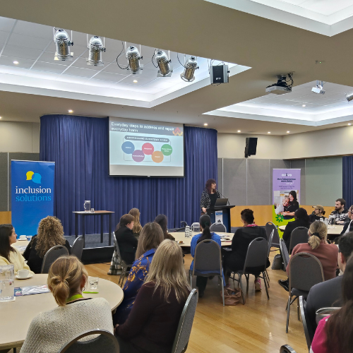 A diverse group of people seated at tables in a well-lit room, engaged in conversation and collaboration. A person is standing in front of everyone and she is having a presentation.