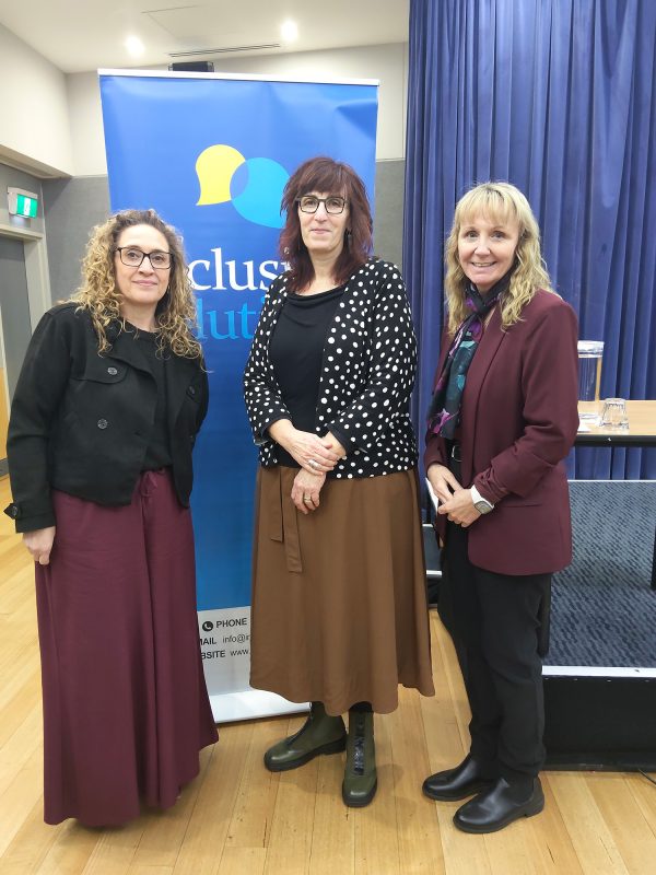 Three women stand together in front of a blue banner with the Inclusion Solutions logotype on it. They are all smiling.