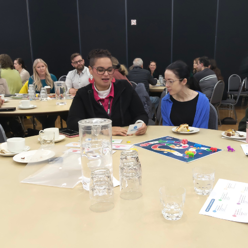 A diverse group of people engaged in discussion while seated around a table. They are playing the Everyday Harm board game.